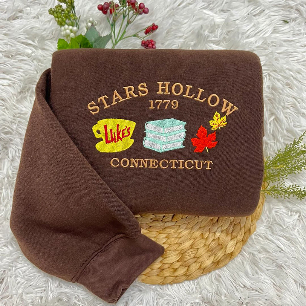 A brown sweatshirt with the 'Stars Hollow 1779' design, featuring embroidered elements including a coffee cup and a diner sign, displayed on a white fluffy surface.