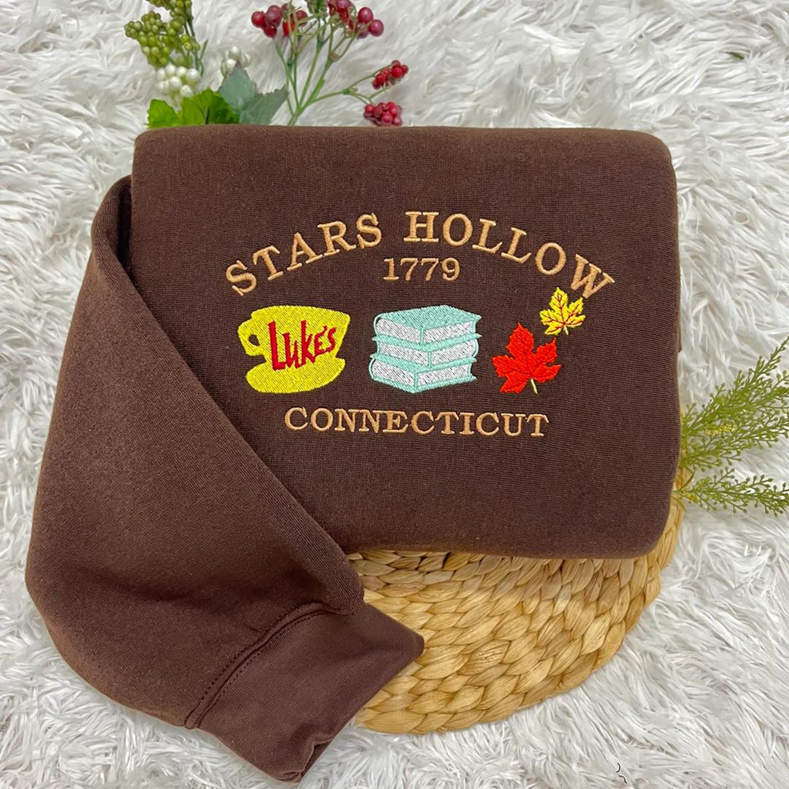 A brown sweatshirt with the 'Stars Hollow 1779' design, featuring embroidered elements including a coffee cup and a diner sign, displayed on a white fluffy surface.