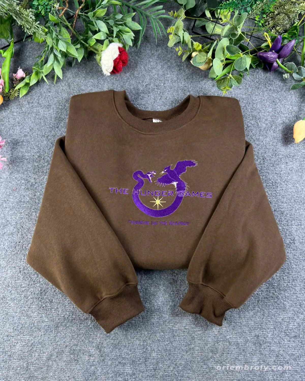 Flat lay of a chocolate brown crewneck sweatshirt embroidered with purple Sunrise on the Reaping Hunger Games logo