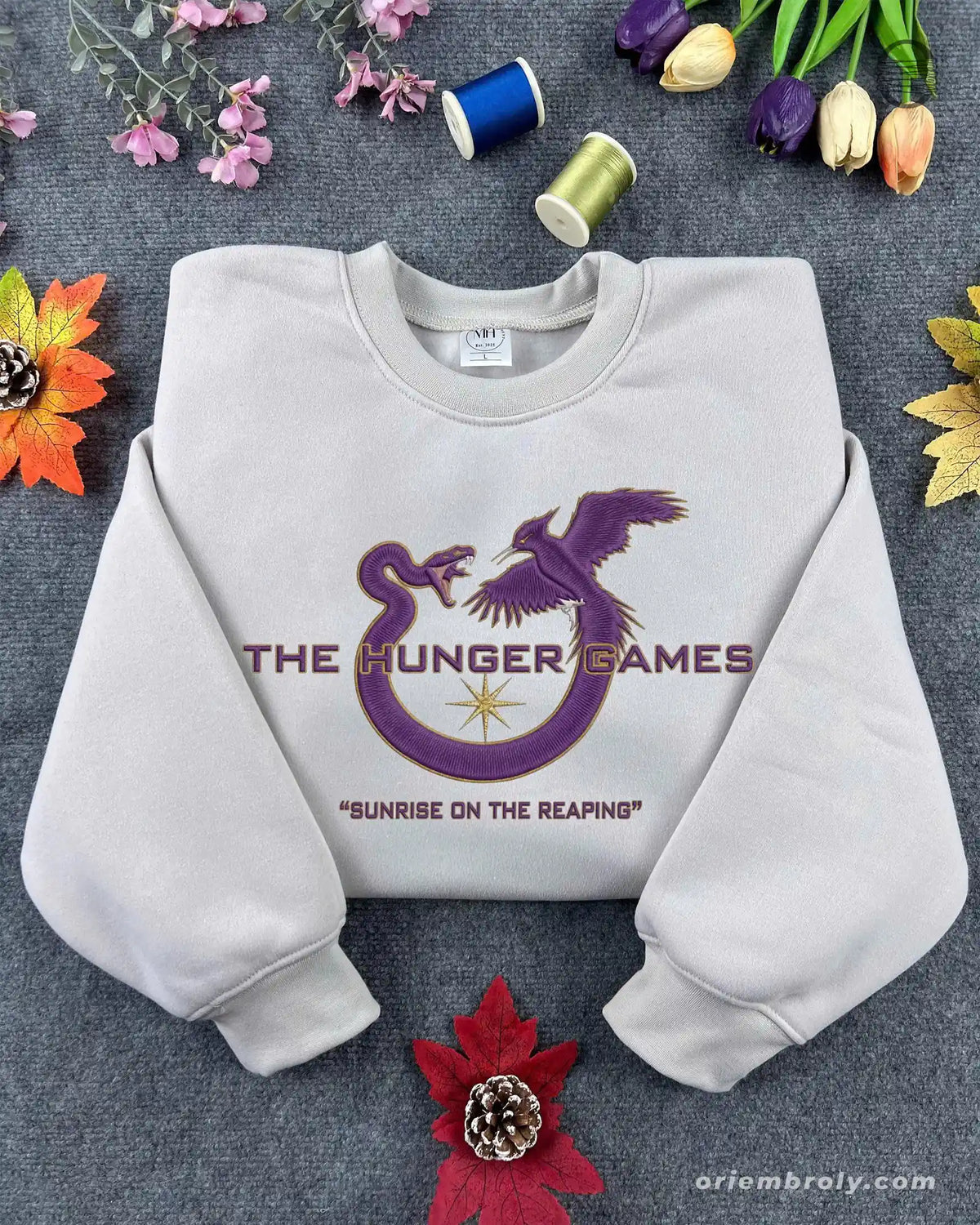Flat lay of a sand color crewneck sweatshirt embroidered with purple Mockingjay logo and Sunrise on the Reaping text