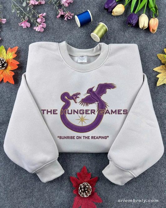 Flat lay of a sand color crewneck sweatshirt embroidered with purple Mockingjay logo and Sunrise on the Reaping text