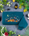 Flatlay view of a dark teal crewneck sweatshirt embroidered with dancing skeleton cats, pumpkins, crescent moon, and yellow stars for Halloween.