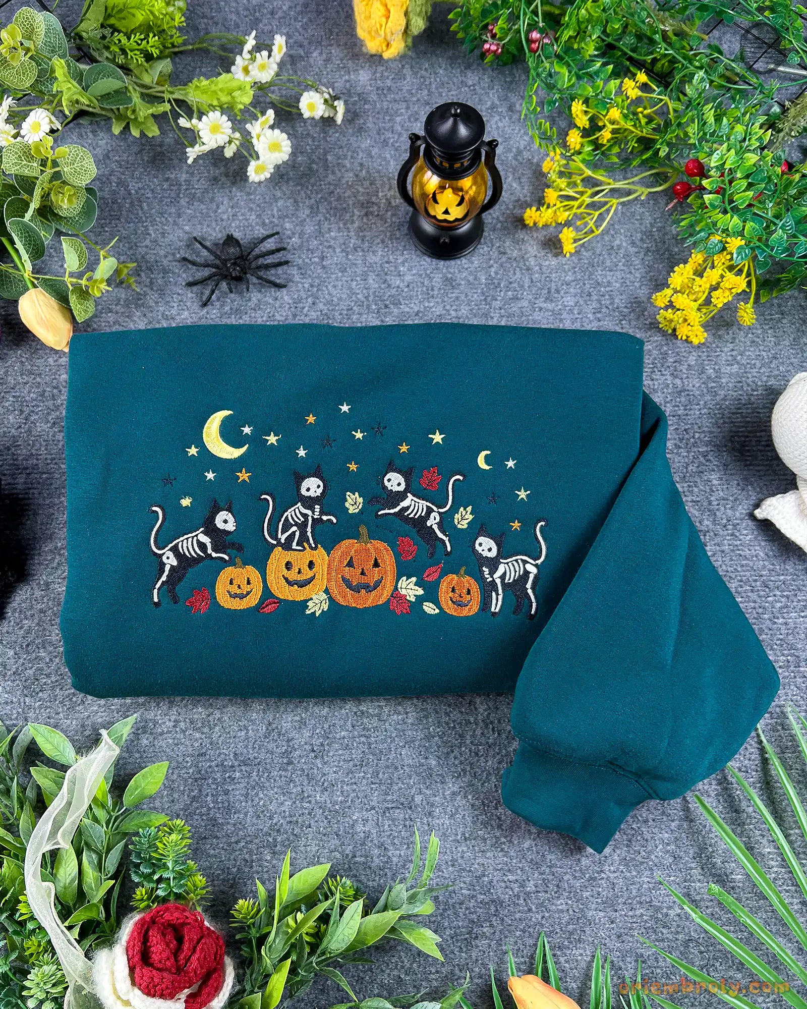 Flatlay view of a dark teal crewneck sweatshirt embroidered with dancing skeleton cats, pumpkins, crescent moon, and yellow stars for Halloween.