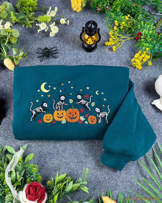 Flatlay view of a dark teal crewneck sweatshirt embroidered with dancing skeleton cats, pumpkins, crescent moon, and yellow stars for Halloween.
