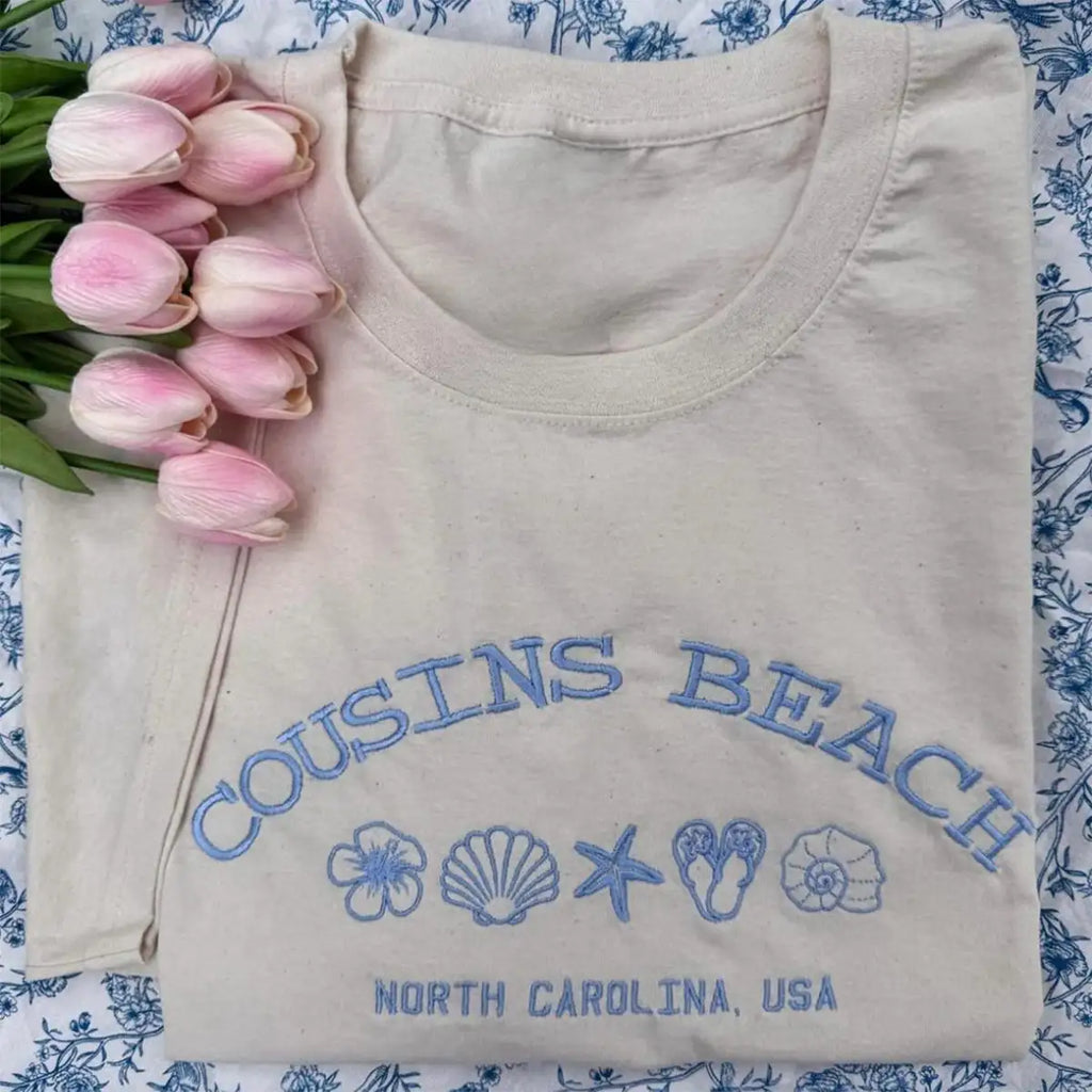 Folded beige shirt with 'Cousins Beach, North Carolina, USA' text and graphics on a floral background.