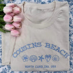 Folded beige shirt with 'Cousins Beach, North Carolina, USA' text and graphics on a floral background.