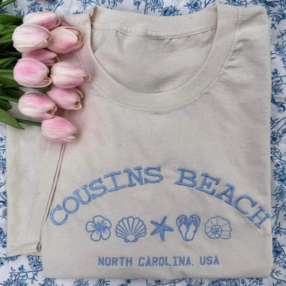 Folded beige shirt with 'Cousins Beach, North Carolina, USA' text and graphics on a floral background.