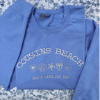 Blue sweatshirt with 'Cousins Beach, North Carolina, USA' text and beach-themed graphics on a floral background.