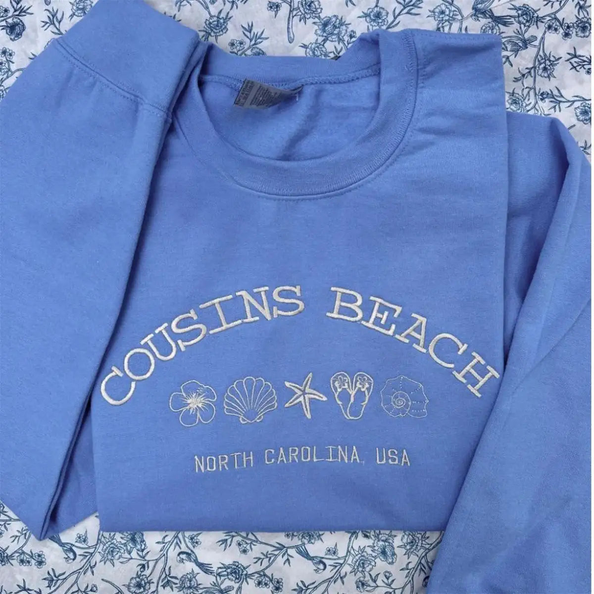 Blue sweatshirt with 'Cousins Beach, North Carolina, USA' text and beach-themed graphics on a floral background.