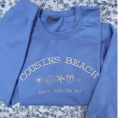 Blue sweatshirt with 'Cousins Beach, North Carolina, USA' text and beach-themed graphics on a floral background.