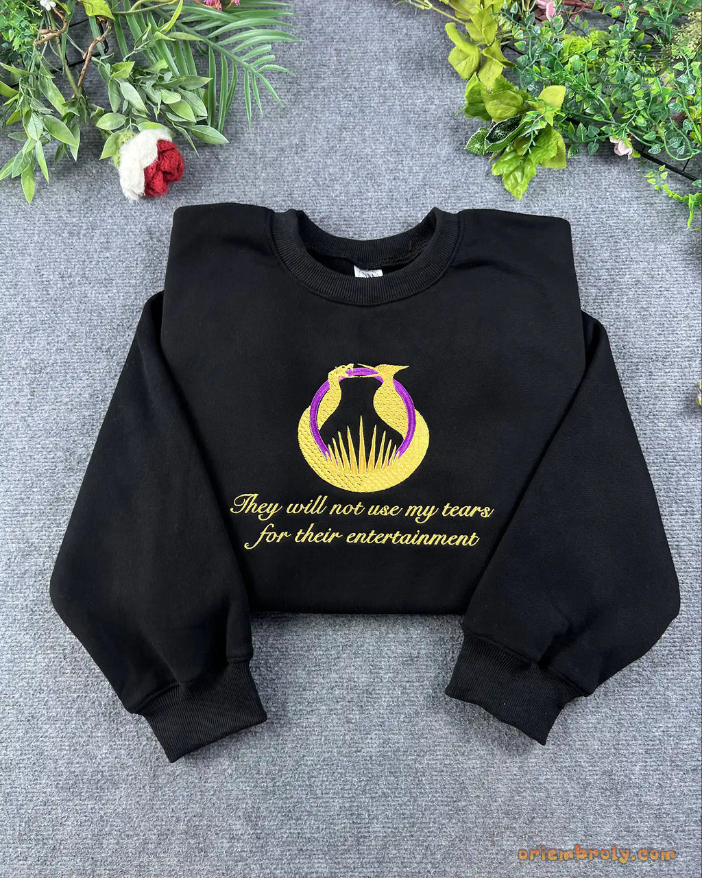 Flat lay of a black crewneck sweatshirt embroidered with gold Hunger Games logo and They will not see my tears quote