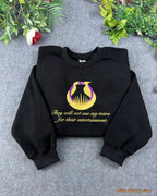 Flat lay of a black crewneck sweatshirt embroidered with gold Hunger Games logo and They will not see my tears quote