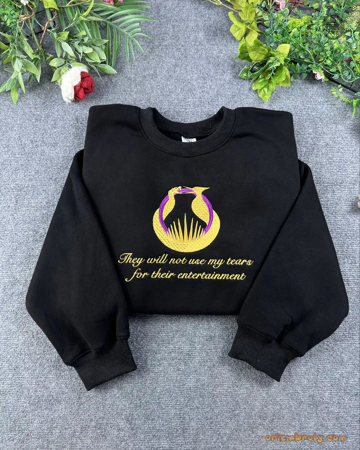 Flat lay of a black crewneck sweatshirt embroidered with gold Hunger Games logo and They will not see my tears quote