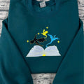 Teal hoodie with embroidered design of a book, cat, and bird on a wooden surface