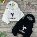 Two hoodies with dragon designs and text on a wooden surface with flowers.