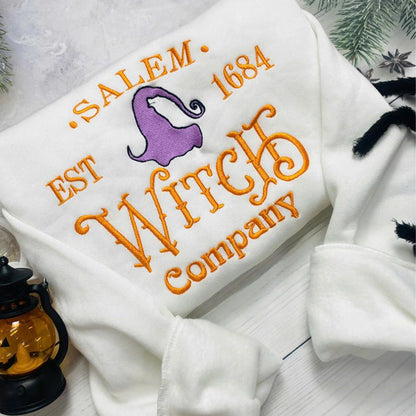 White sweatshirt with embroidered text and witch hat design on a light background.