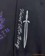 Close-up of silver dagger and Violence quote embroidered on the left sleeve of a black Basgiath War College sweatshirt