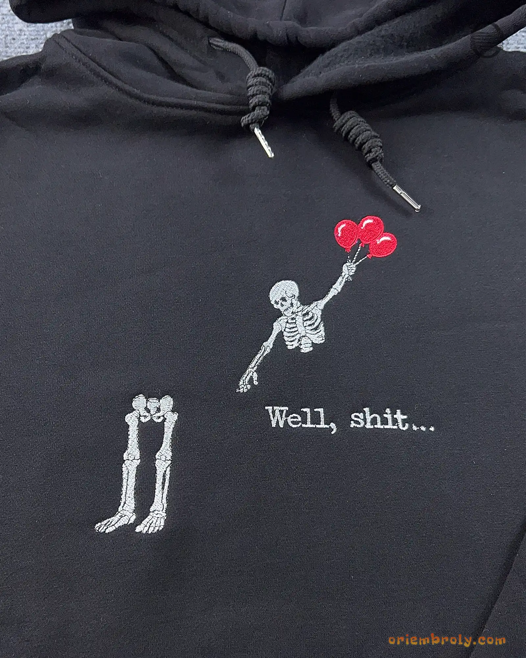 Close-up of Well shit embroidered text with a skeleton flying with red balloons leaving its legs on a black hoodie