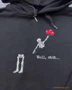 Close-up of Well shit embroidered text with a skeleton flying with red balloons leaving its legs on a black hoodie