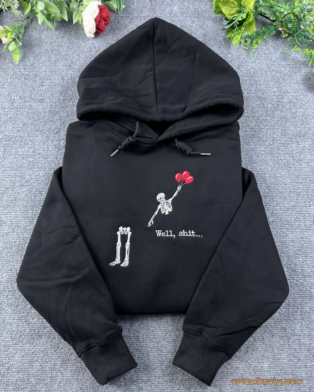 Flat lay of a black hoodie embroidered with a skeleton floating away with red balloons leaving legs behind and Well shit text