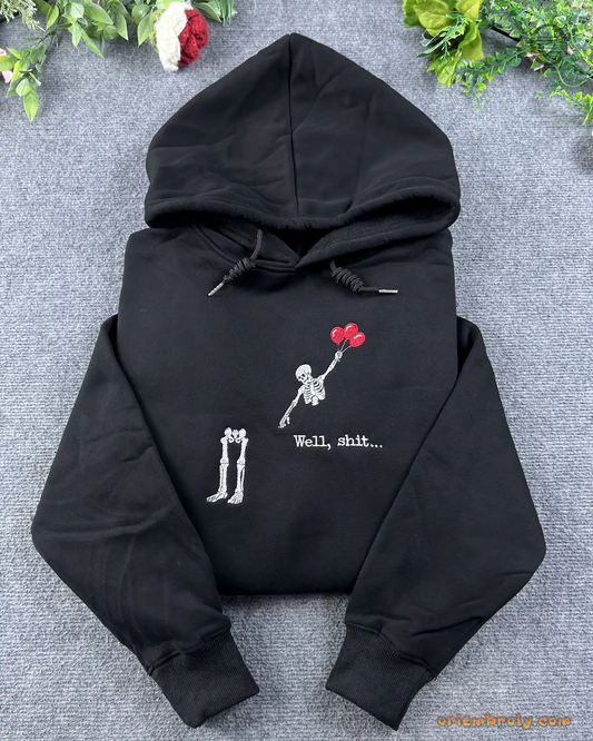 Flat lay of a black hoodie embroidered with a skeleton floating away with red balloons leaving legs behind and Well shit text