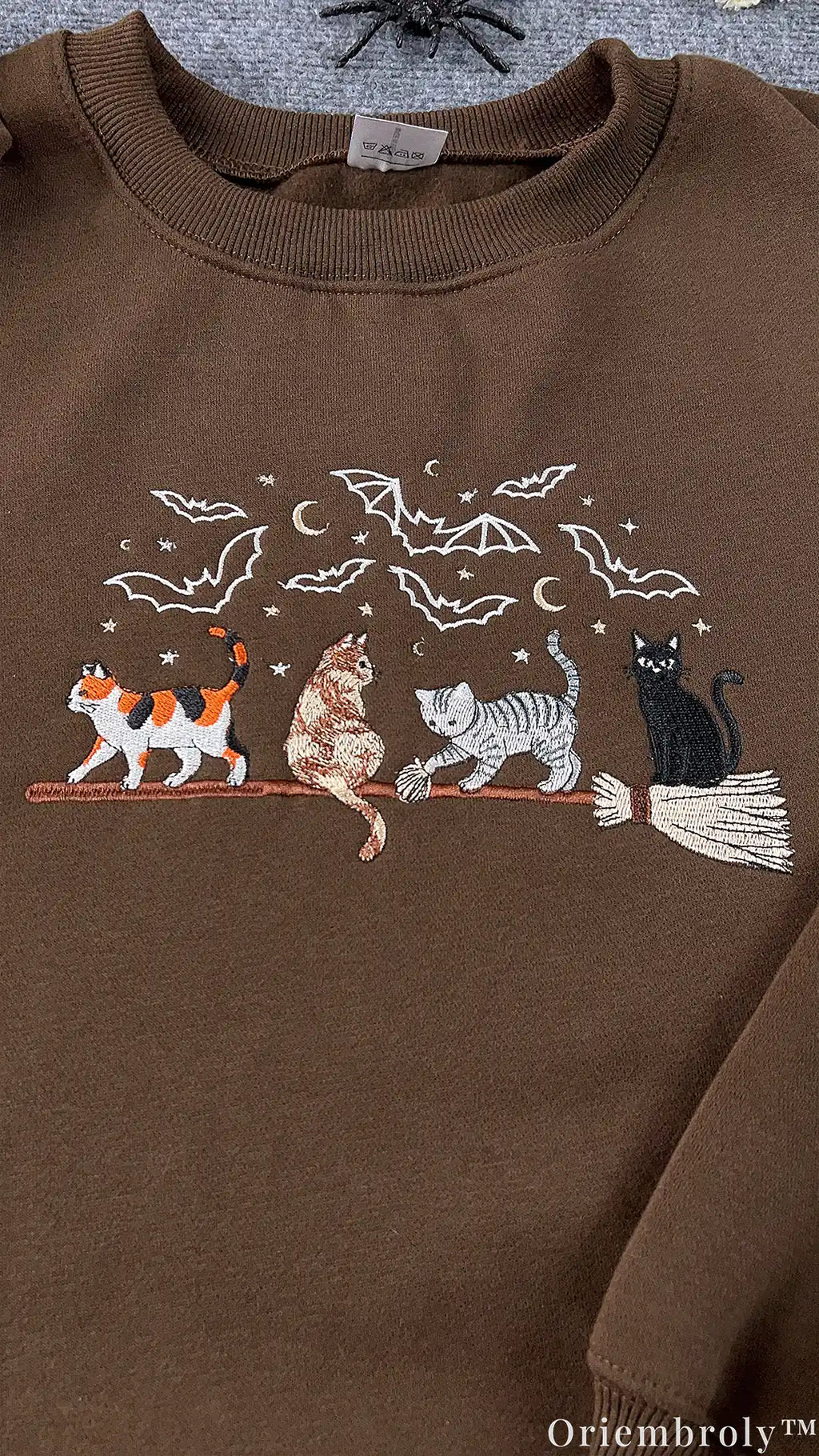 Detailed view of embroidered calico and black cats on a magic broomstick on a chocolate brown sweatshirt