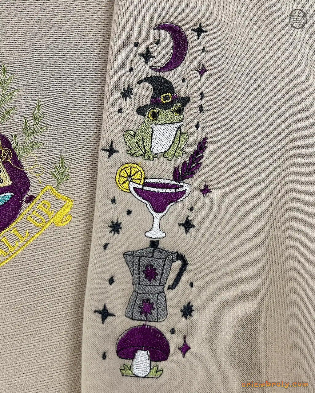Detailed view of witchy frog, cocktail, and purple mushroom sleeve embroidery on a beige crewneck sweater