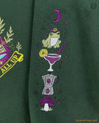 Detailed view of witchy frog, cocktail glass, and mushroom sleeve embroidery on a forest green crewneck