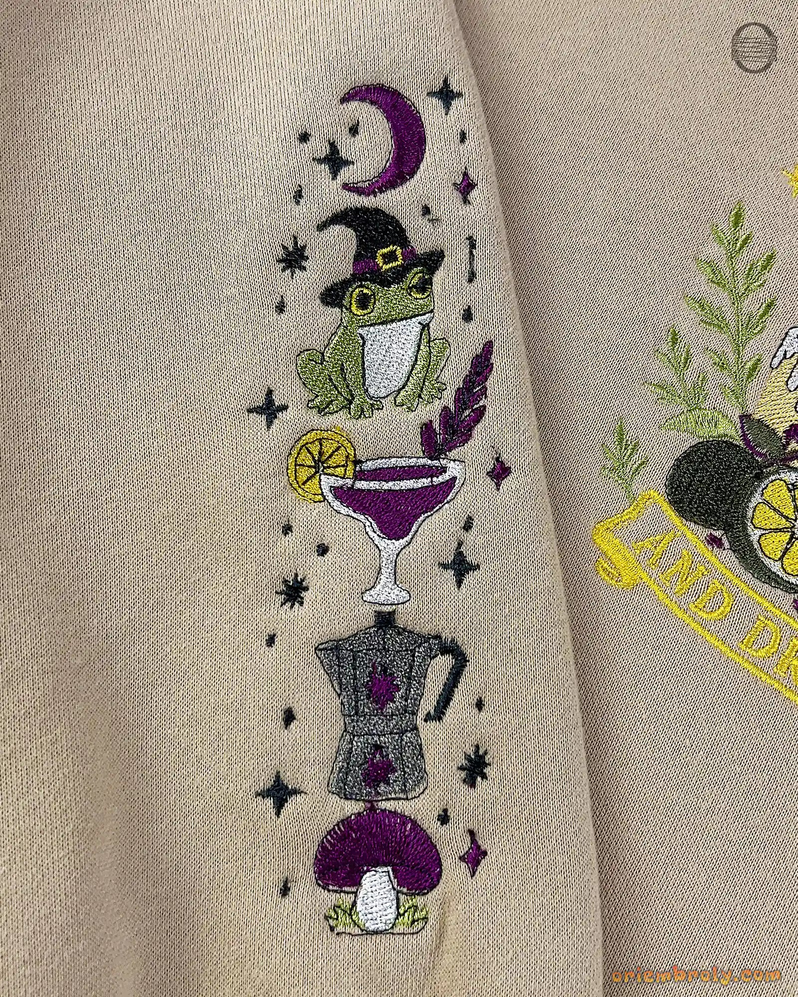 Close-up of sleeve embroidery featuring a wizard frog in hat, cocktail glass, and moka pot on a beige sweatshirt