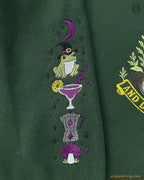 Close-up of wizard frog wearing hat and margarita cocktail sleeve embroidery on a forest green sweater