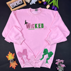 Pink sweatshirt with 'WICKED' text and decorative elements on a dark surface with flowers and a calendar.