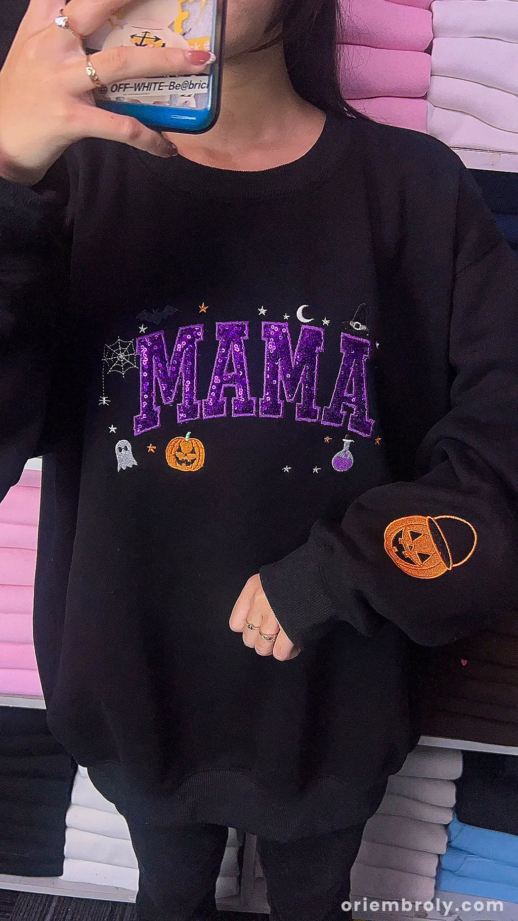 Model wearing an oversized black Mama Halloween sweatshirt with purple sequin inner text and pumpkin sleeve print.
