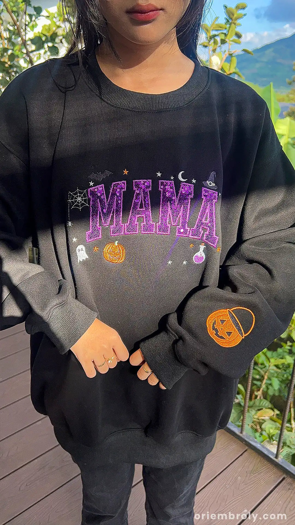 Model presenting a black Mama Halloween sweatshirt with purple sequin lettering and pumpkin sleeve print outdoors.