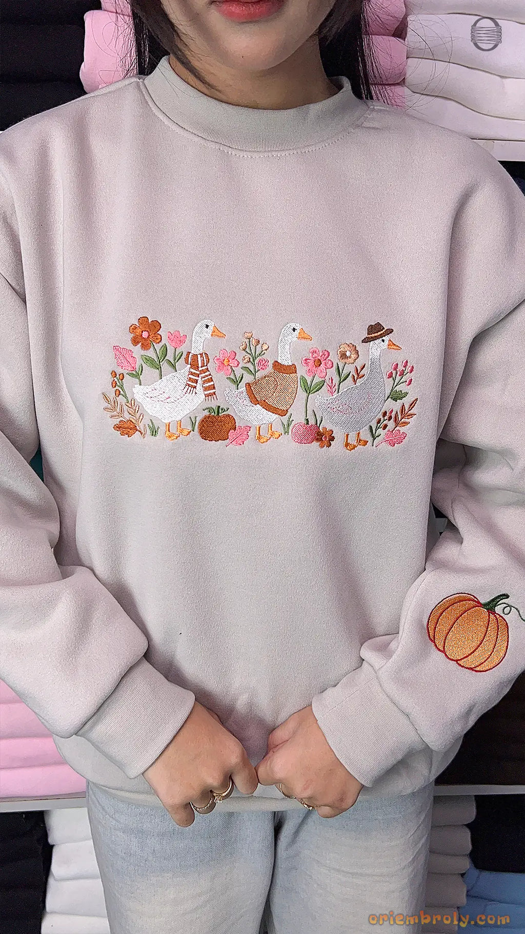 Model wearing an oversized light gray sweatshirt with embroidered ducks on the chest and a pumpkin print on the sleeve indoors.