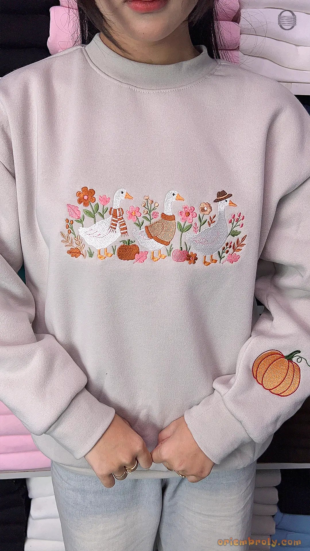 Model wearing an oversized light gray sweatshirt with embroidered ducks on the chest and a pumpkin print on the sleeve indoors.