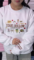 Model wearing a white Stars Hollow Connecticut embroidered crewneck sweatshirt with sleeve detail indoors.