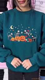 Model wearing an oversized dark teal Halloween sweatshirt featuring embroidered skeleton cats and pumpkins indoors.