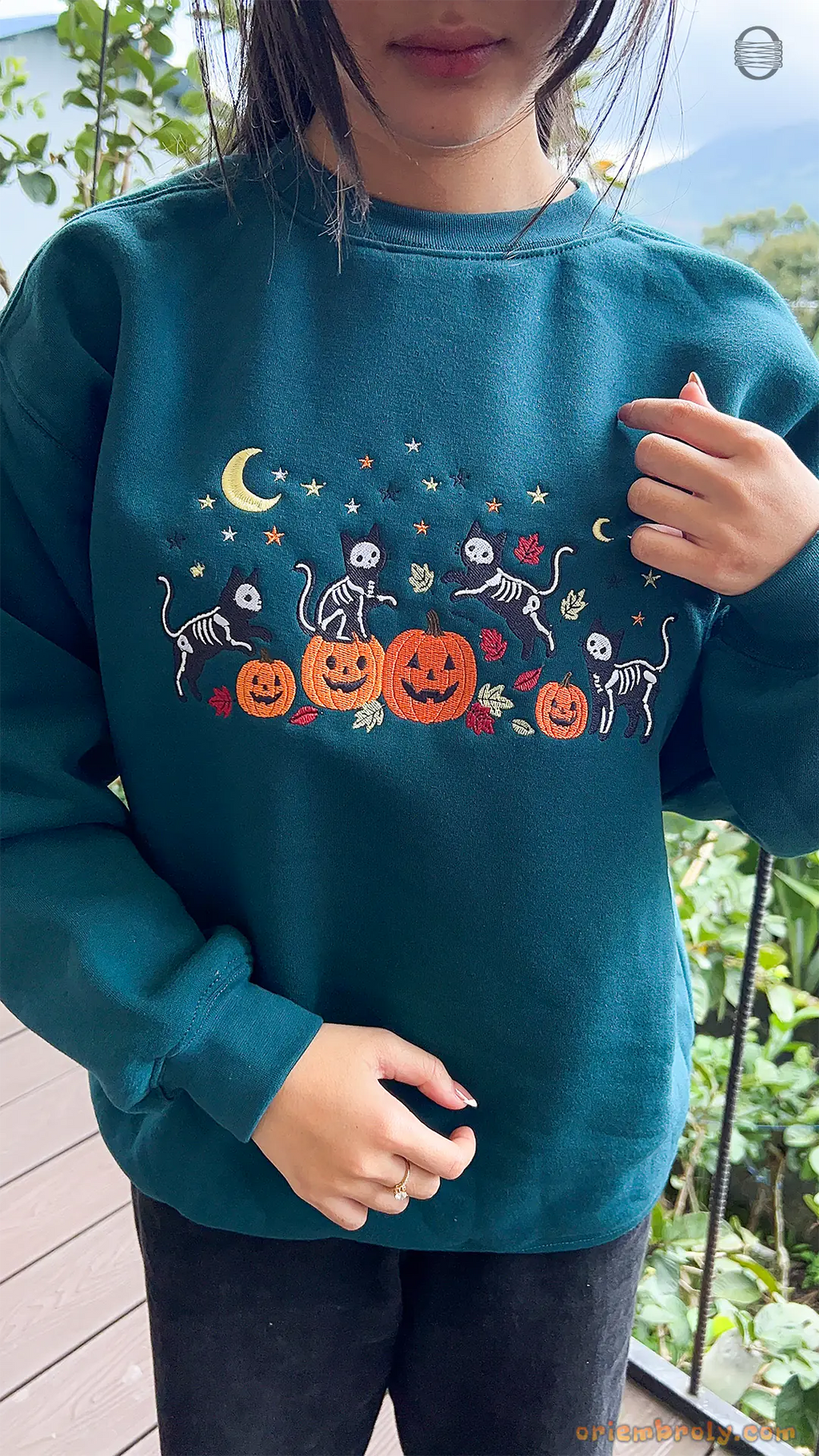 Model showcasing a dark teal skeleton cats and pumpkin embroidered Halloween sweater in an outdoor fall setting