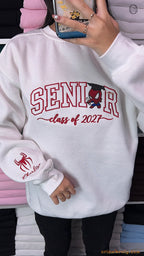Model wearing a white Senior Class of 2027 embroidered crewneck sweatshirt with Spider-Man sleeve detail indoors.