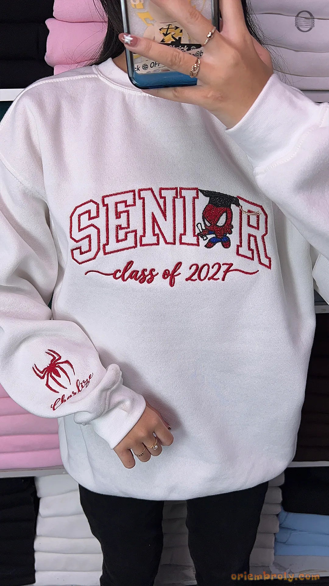 Model wearing a white Senior Class of 2027 embroidered crewneck sweatshirt with Spider-Man sleeve detail indoors.